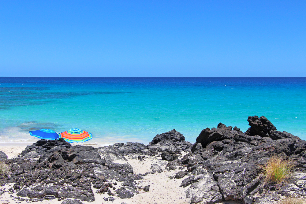 Manini'owali Beach in Kua Bay, Big Island, Hawaii. The view on the two colorful beach umbrellas behind black lava rock with beautiful turquoise sea and white sand in the background.